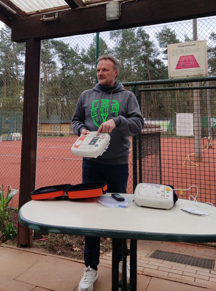 Volker Beckmann vom TC Metelen 64 bei der Einweisung in die Nutzung des Defibrillator. Er zeigt auf die Symbole des Gerätes, im Hintergrund der rote Ascheplatz des Tennisvereins.
