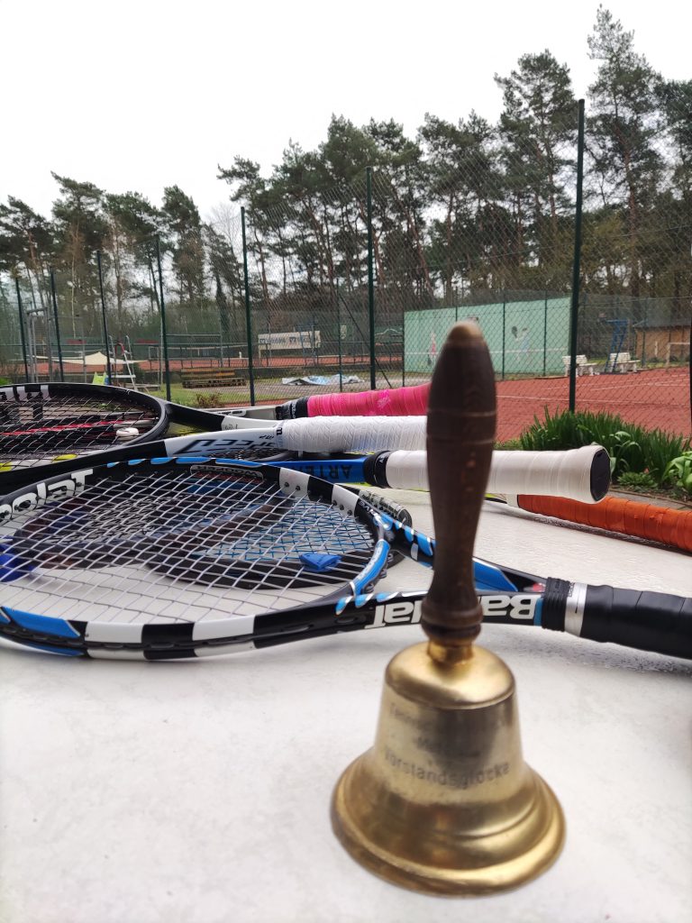 Zu sehen ist die Vorstandsglocke des Tennisvereins Metelen. Sie steht auf einem Tisch, dahinter liegen mehrere Tennisschläger übereiander und warten auf ihren Einsatz.